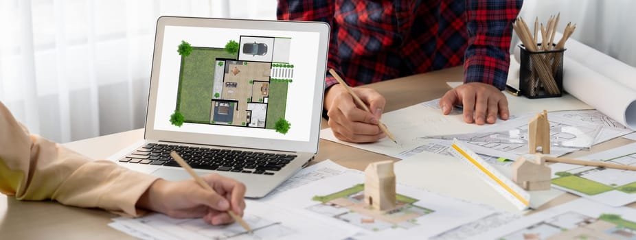 Professional architect represented ECO house displayed on laptop to project manager on messy table with wooden block, blueprint, architectural document placed at modern office. Close up. Delineation.