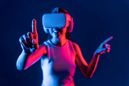 Smart female stand with surrounded by cyberpunk neon light wear VR headset connecting metaverse, futuristic cyberspace community technology. Woman using finger pointing virtual object. Hallucination.