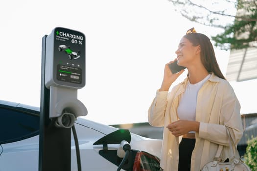 Young woman holding shopping bag talking on the phone while recharging EV car battery from charging station at city mall parking lot. Modern woman go shopping by eco car. Expedient