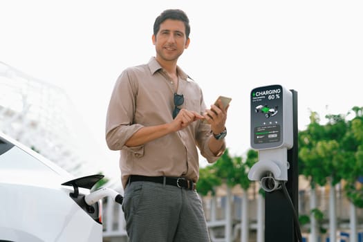 Young man use smartphone to pay for electricity at public EV car charging station green city park. Modern environmental and sustainable urban lifestyle with EV vehicle. Expedient