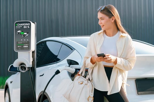 Young woman holding shopping bag and use smartphone to pay for electricity for recharging EV car battery from charging station at city mall parking lot. Modern woman go shopping by eco car. Expedient