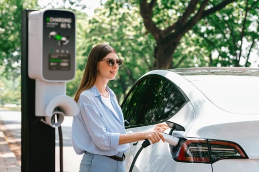 Young woman recharging battery for electric car during road trip travel EV car in natural forest or national park. Eco friendly travel during vacation and holiday. Exalt