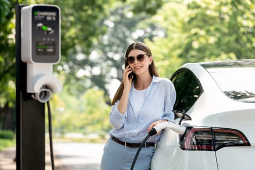 Young woman talking on smartphone while recharging electric car battery charging from EV charging station during vacation holiday road trip at national park or summer forest. Exalt