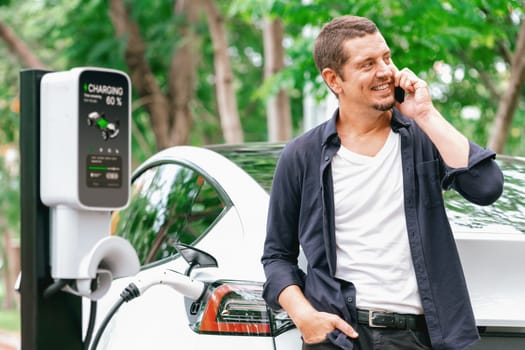 Man talking on smartphone while recharging electric car battery charging from EV charging station during vacation holiday road trip at national park or summer forest. Exalt