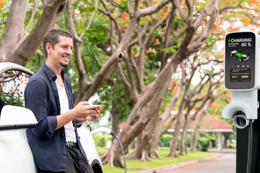 Man using smartphone online banking application to pay for electric car battery charging from EV charging station during autumn vacation holiday trip at national park or autumnal forest. Exalt