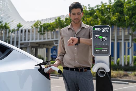 Young man checking time on smartwatch while EV charger to recharging battery from charging station in city mall parking lot. Rechargeable EV car for sustainable eco friendly urban travel. Expedient