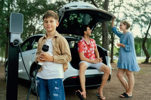 Little boy holding EV charger and point at camera with his family sitting on the trunk in background. Road trip travel with alternative energy charging station for eco-friendly car concept. Perpetual