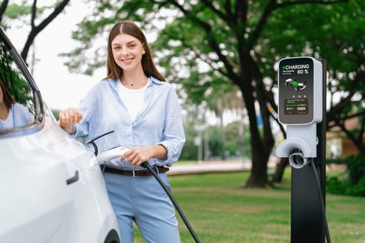 Young woman recharge EV electric vehicle's battery from EV charging station in outdoor green city park scenic. Eco friendly urban transport and commute with eco friendly EV car travel. Exalt