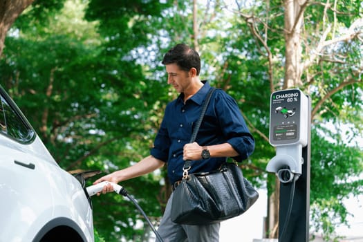 Young man recharge electric car's battery from charging station in outdoor green city park in springtime. Rechargeable EV car for sustainable environmental friendly urban travel lifestyle. Expedient