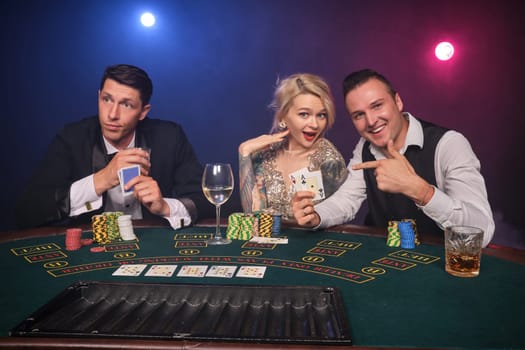 Two athletic male and alluring female are playing poker at casino. Youth are making bets waiting for a big win. They are smiling and posing sitting at the table against a red and blue backlights on black smoke background. Cards, chips, money, gambling, entertainment concept.