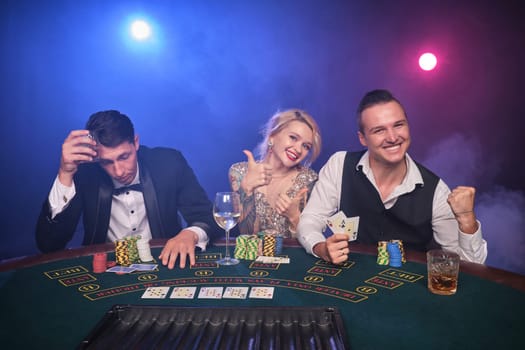 Two athletic men and alluring woman are playing poker at casino. Youth are making bets waiting for a big win. They are smiling and posing sitting at the table against a red and blue backlights on black smoke background. Cards, chips, money, gambling, entertainment concept.