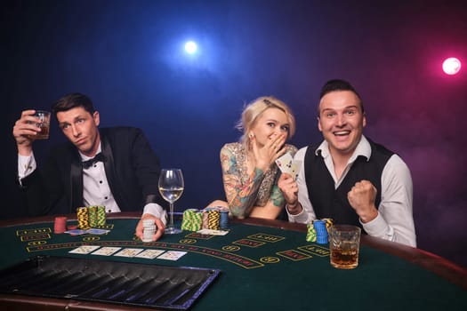 Two elegant fellows and attractive lady are playing poker at casino. Youth are making bets waiting for a big win. They are smiling and posing sitting at the table against a red and blue backlights on black smoke background. Cards, chips, money, gambling, entertainment concept.