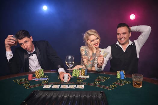 Two elegant men and attractive maiden are playing poker at casino. Youth are making bets waiting for a big win. They are smiling and looking at the camera while sitting at the table against a red and blue backlights on black smoke background. Cards, chips, money, gambling, entertainment concept.