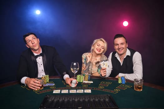 Two elegant guys and attractive girl are playing poker at casino. Youth are making bets waiting for a big win. They are smiling and posing sitting at the table against a red and blue backlights on black smoke background. Cards, chips, money, gambling, entertainment concept.