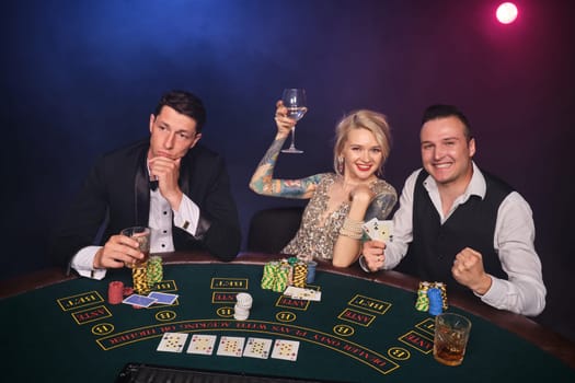 Two elegant men and attractive woman are playing poker at casino. Youth are making bets waiting for a big win. They are smiling and drinking alcohol while sitting at the table against a red and blue backlights on black smoke background. Cards, chips, money, gambling, entertainment concept.