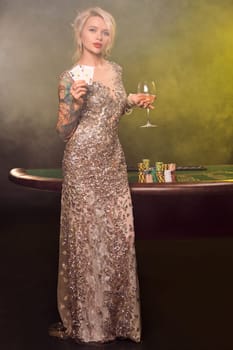Full length shot of a lovely woman with blond hair, tattoed hands and perfect make-up, dressed in a silver shiny dress. She is standing against a gambling table, with two playing cards in her hands and smiling. Poker concept on a black smoke background with yellow and white backlights. Casino.