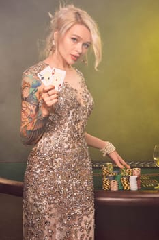Close-up shot of a cute female with blond hair, tattoed hands and perfect make-up, dressed in a silver shiny dress. She is standing against a gambling table, with two playing cards in her hands and looking at you. Poker concept on a black smoke background with yellow and white backlights. Casino.
