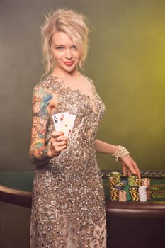 Close-up shot of a cute woman with blond hair, tattoed hands and perfect make-up, dressed in a silver shiny dress. She is standing against a gambling table, with two playing cards in her hands and smiling. Poker concept on a black smoke background with yellow and white backlights. Casino.