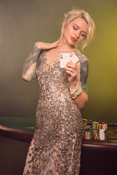 Close-up shot of a pretty girl with blond hair, tattoed hands and perfect make-up, dressed in a silver shiny dress. She is standing against a gambling table, with two playing cards in her hands and looking at it. Poker concept on a black smoke background with yellow and white backlights. Casino.