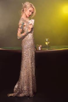 Full length shot of a pretty lady with blond hair, tattoed hands and perfect make-up, dressed in a silver shiny dress. She is standing sideways against a gambling table, with two playing cards in her hands and smiling. Poker concept on a black smoke background with yellow and white backlights. Casino.