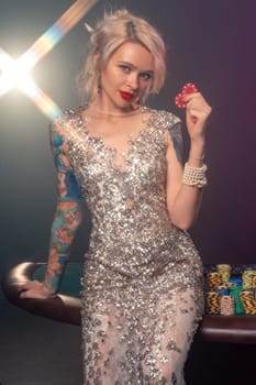 Close-up shot of an graceful lady with blond hair, tattoed hands and perfect make-up, dressed in a silver shiny dress. She is standing against a gambling table, with red chips in her hands and smiling. Poker concept on a black smoke background with a backlight. Casino.