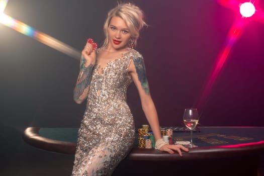 Medium close-up shot of a charming lady with blond hair, tattoed hands and perfect make-up, dressed in a silver shiny dress. She is standing sideways against a gambling table, with red chips in her hands and smiling. Poker concept on a black smoke background with pink and blue backlights. Casino.