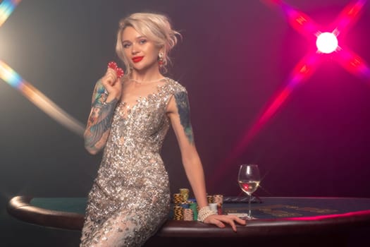 Medium close-up shot of a charming girl with blond hair, tattoed hands and perfect make-up, dressed in a silver shiny dress. She is standing against a gambling table, with red chips in her hands and smiling. Poker concept on a black smoke background with pink and blue backlights. Casino.