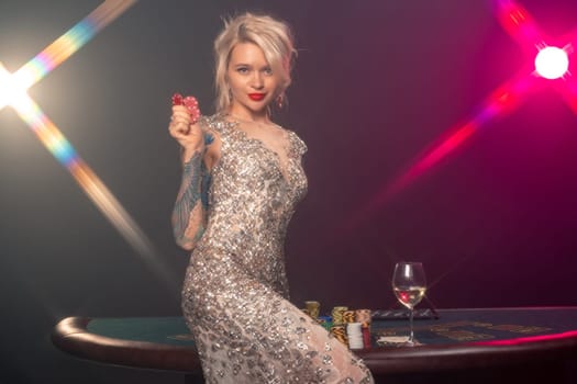 Medium close-up shot of a charming female with blond hair, tattoed hands and perfect make-up, dressed in a silver shiny dress. She is sitting on a gambling table, with red chips in her hands and looking at the camera. Poker concept on a black smoke background with pink and blue backlights. Casino.