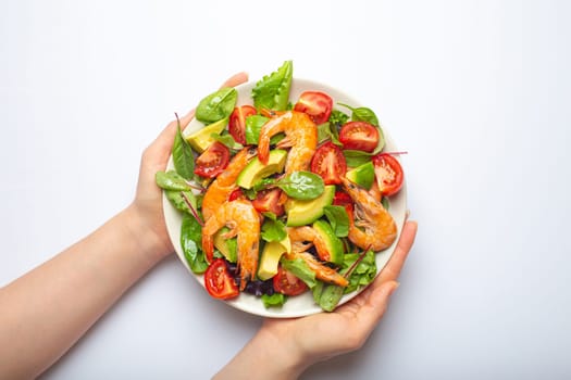 Female hands holding healthy salad with grilled shrimps, avocado, cherry tomatoes and green leaves on white plate isolated on white background top view. Clean eating, nutrition and dieting concept..