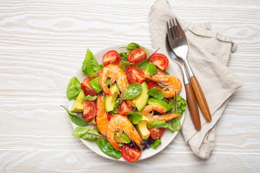 Healthy salad with grilled shrimps, avocado, cherry tomatoes and green leaves on white plate with cutlery on white wooden rustic background top view. Clean eating, nutrition and dieting concept..