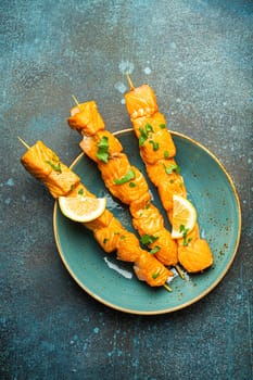 Grilled barbecue salmon skewers seasoned with green parsley and lemon on ceramic plate on dark blue concrete rustic table background top view, healthy eating.