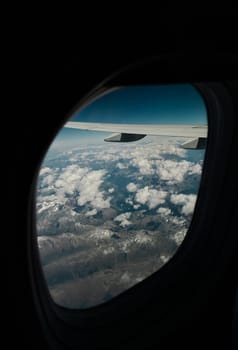 A beautiful view of an airplane wing, with rocky mountain tops in white clouds through the porthole window, close-up side view.