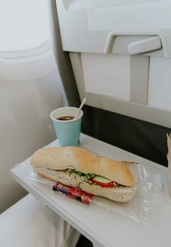 One appetizing sandwich with tuna, tomatoes and a paper glass of tea lie on a table on the plane, close-up side view.