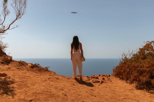 One beautiful young brunette girl in a white robe stands from her back on the edge of a cliff, looking at the blue sea below, and a drone flying in the sky films her with a camera, close-up side view.