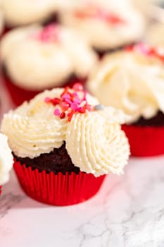 Piping white chocolate ganache frosting on top of red velvet cupcakes and topping with sprinkles.