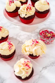 Freshly baked velvet cupcakes with white chocolate ganache frosting decorated with sprinkles.