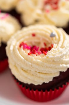 Freshly baked velvet cupcakes with white chocolate ganache frosting decorated with sprinkles.