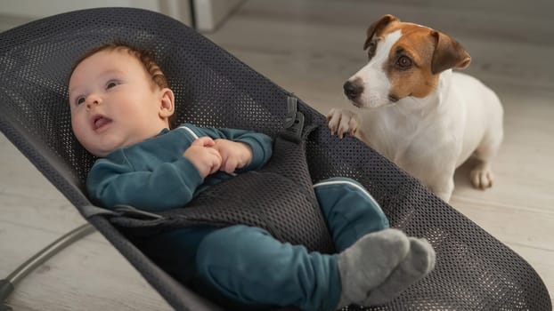 A dog sits next to a cute three-month-old boy dressed in a blue overalls in a baby lounger