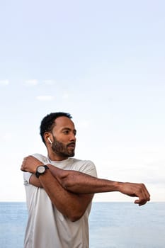 Vertical portrait of African American man stretching arm, warming up before workout and running session outdoors. Male exercising near the ocean. Health and fitness concept.