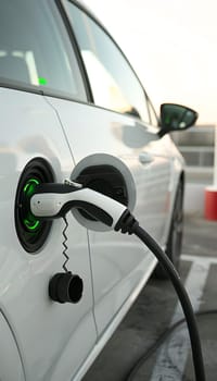 An electric vehicle is being charged at a charging station, its sideview mirror reflecting the automotive lighting while the alloy wheels rest on the ground