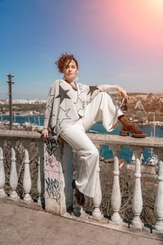 Woman walks around the city, lifestyle. A young beautiful woman in white trousers and a sweater sits on a white fence with balusters and overlooks the sea bay and the city