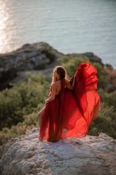 Woman sunset sea red dress, back view a happy beautiful sensual woman in a red long dress posing on a rock high above the sea on sunset