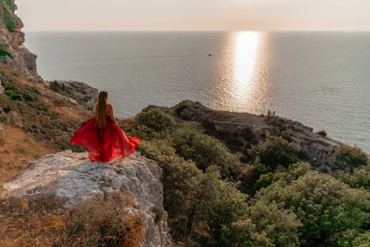 Woman sunset sea red dress, back view a happy beautiful sensual woman in a red long dress posing on a rock high above the sea on sunset