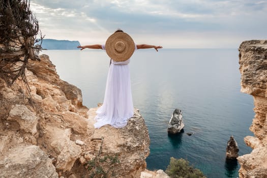 woman white dress stands on a rocky cliff overlooking the ocean. She is wearing a straw hat and she is enjoying the view