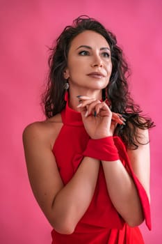 A woman in a red dress is standing in front of a pink background. She is wearing a red scarf and earrings