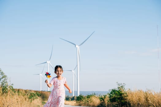 Little girl's playful interaction near windmills holding pinwheels while running. Embracing wind energy education promoting clean electricity and a cheerful childhood.