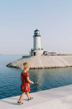 Little girl walks along the embankment overlooking the lighthouse on the breakwater. Back view. High quality photo
