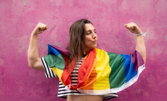 Lesbian woman in strong arm pose with rainbow flag isolated on pink background.