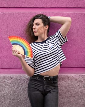 Transgender woman with rainbow fan posing sexy on pink. Concept of pride expressing support for the LGBTQ+ community.