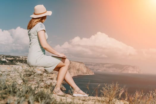 Digital nomad, woman in the hat, a business woman with a laptop sits on the rocks by the sea during sunset, makes a business transaction online from a distance. Freelance, remote work on vacation.